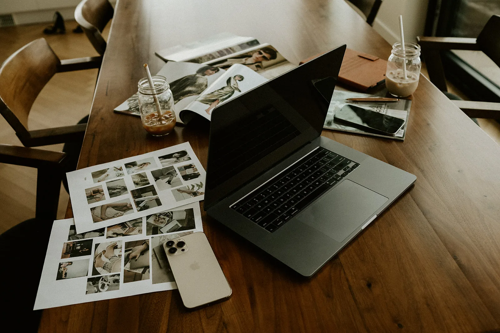 Laptop on a wooden table surrounded by photo layouts and drinks.